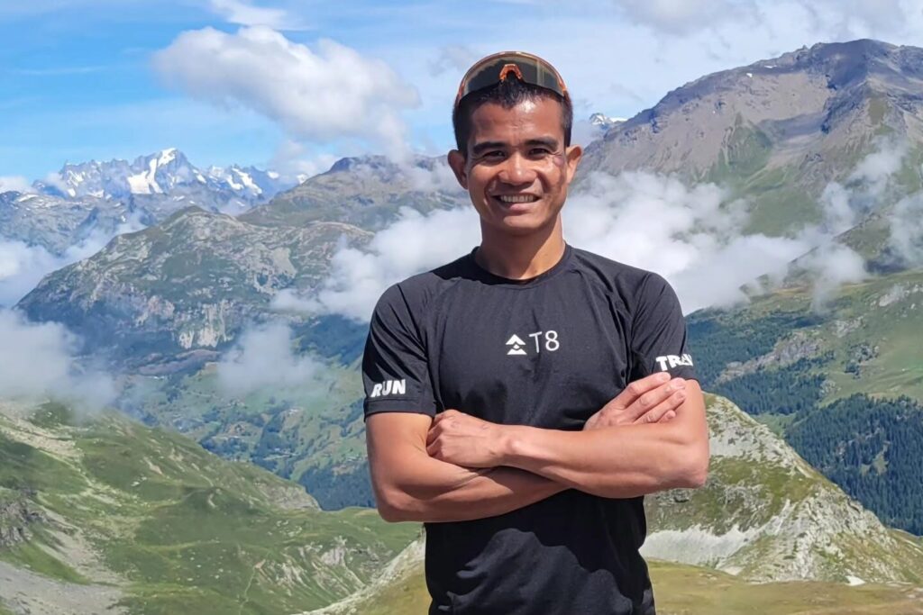 John Ray Onifa in a T8 shirt with arms crossed, Alpine mountains behind him